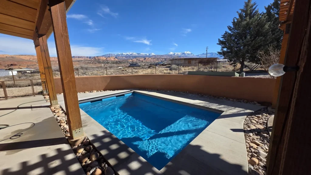 Bright blue fiberglass pool under a timber pergola, with a stucco wall and snow on the La Sal Mountains
