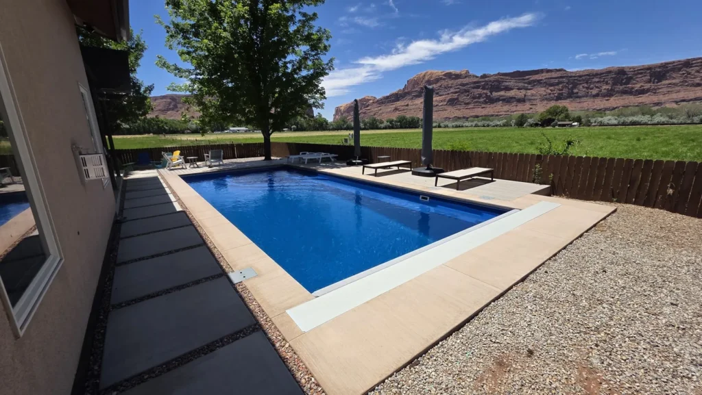 Rectangular blue pool beside a house, with a deck-mounted cover lid, a hay field, and red sandstone cliffs