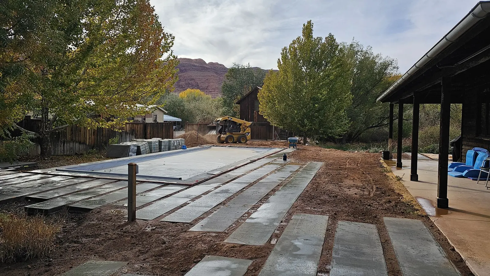 Fiberglass pool mid-install, with the cover on, concrete pavers set in compacted red soil, and a skid steer finishing the grade