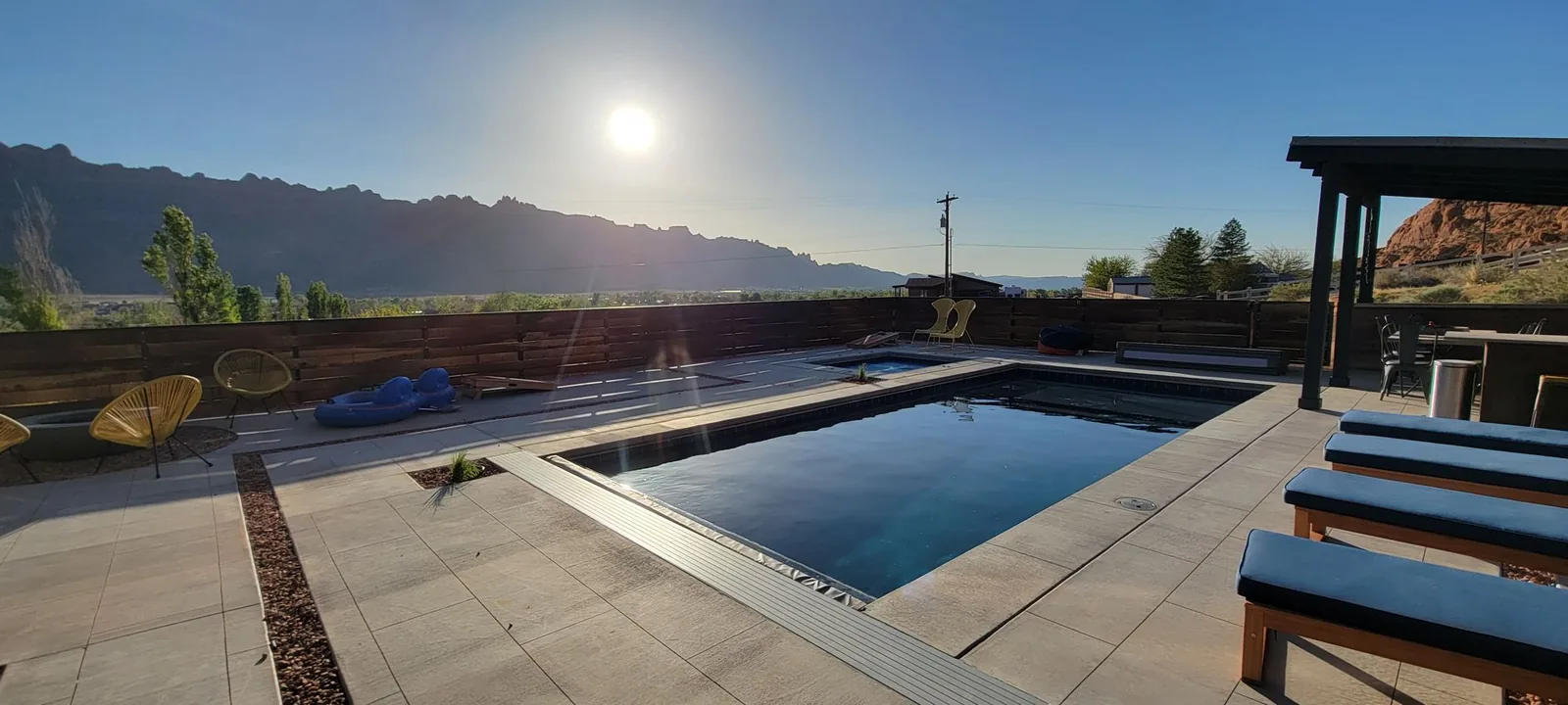 Pool and spa on a tiled hillside patio with lounge chairs, a pergola, and a sandstone ridge in morning sun