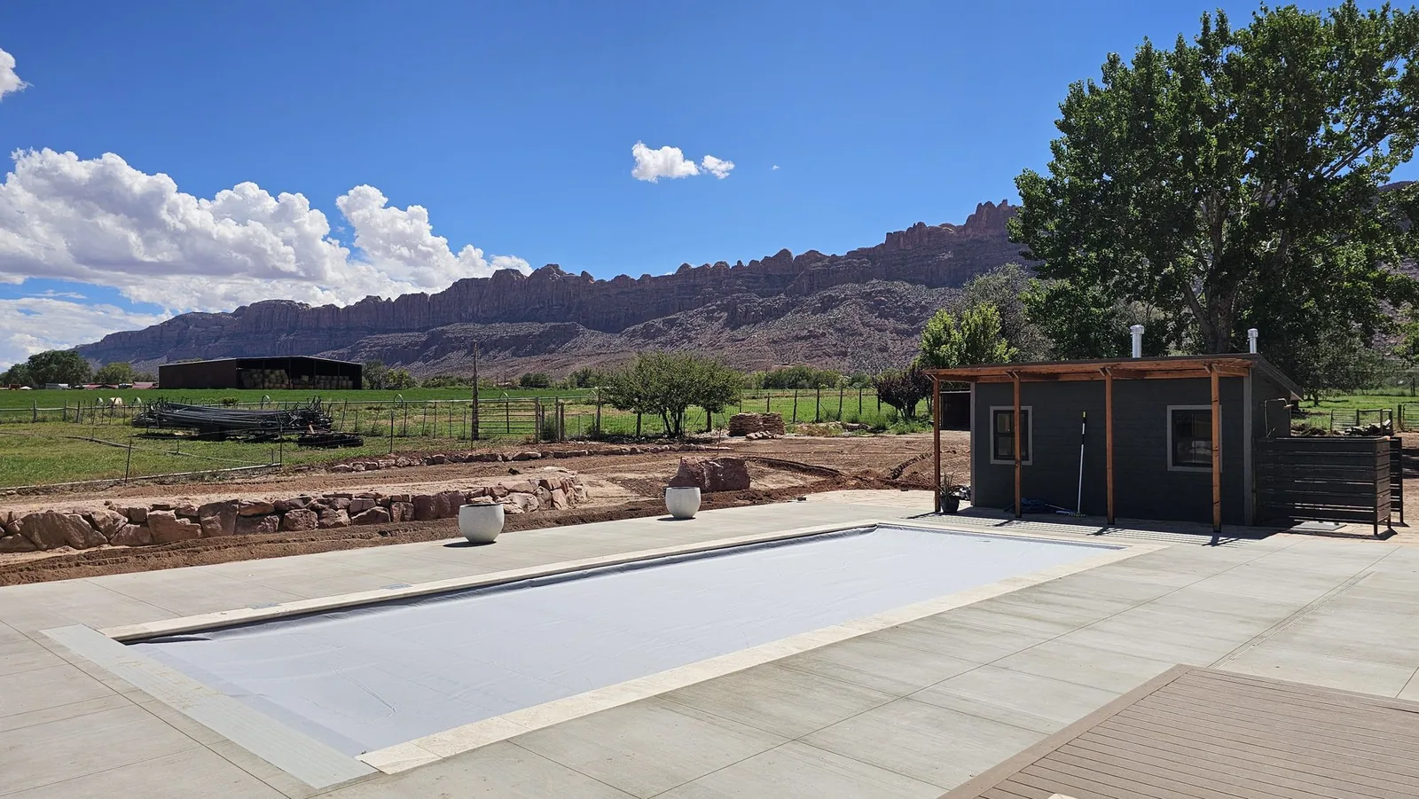 Covered pool on a wide concrete deck beside a pool house, with a pergola, hay barn, and a long red rock ridge