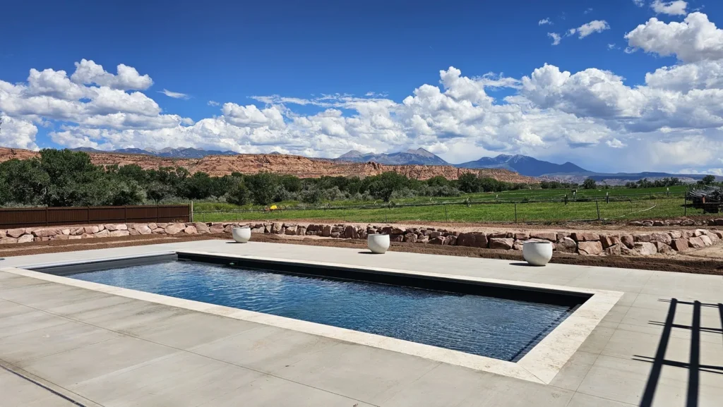 Long dark-finish fiberglass pool with light stone coping, white planters, a sandstone wall, and the La Sal Mountains under summer clouds