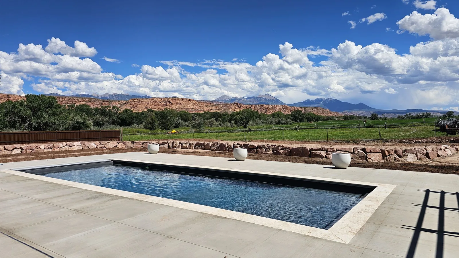 Long dark-finish fiberglass pool with light stone coping, white planters, a sandstone wall, and the La Sal Mountains under summer clouds