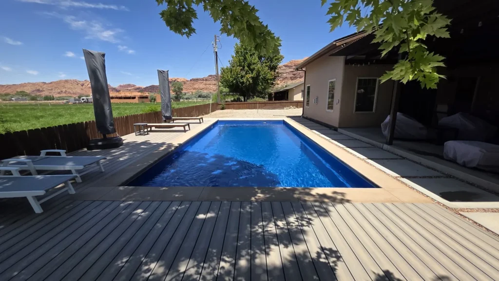 Bright blue fiberglass pool with a composite deck and lounge chairs, a field and red rock beyond